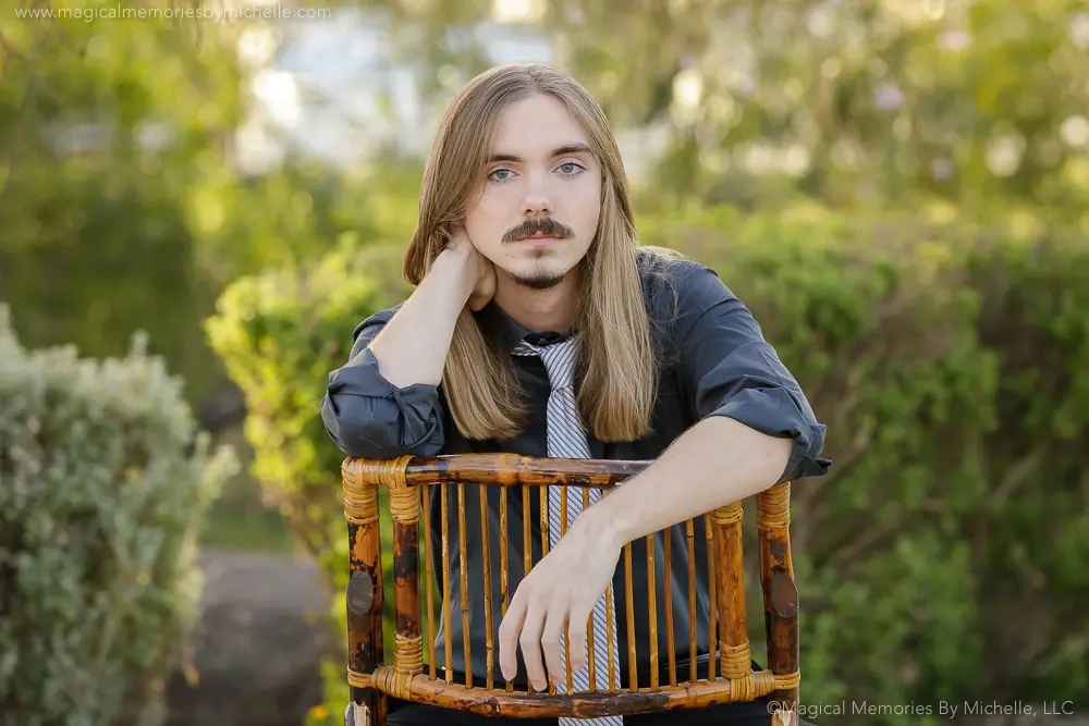 Senior boy leaning on a wooden chair outdoors wearing a black shirt and striped tie for formal senior pictures in Mesa, Arizona.