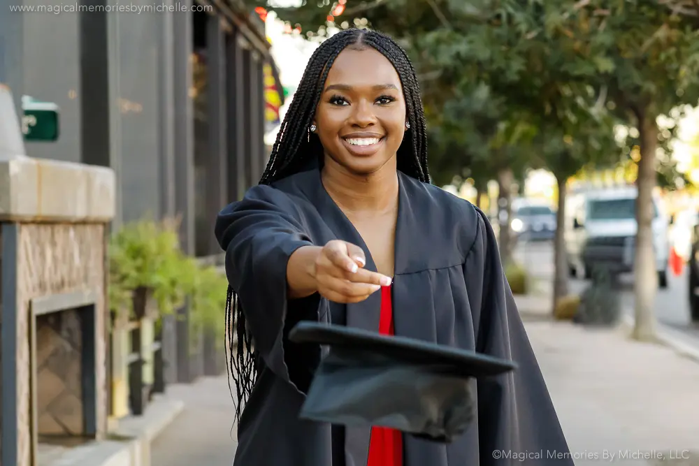 Smiling Black high school senior in cap and gown tosses her graduation cap toward the camera during her senior photo session in Downtown Gilbert, Arizona.