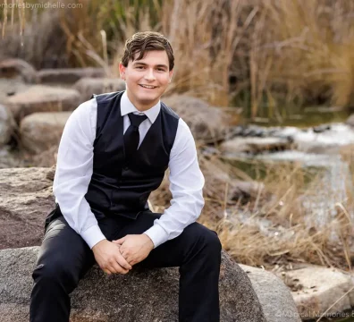 Senior guy sitting on a rock by water wearing a black vest and tie for formal senior portraits in Mesa, Arizona.
