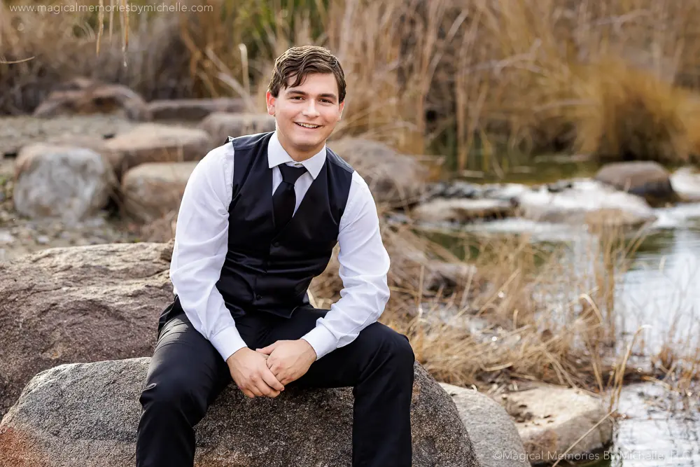 Senior guy sitting on a rock by water wearing a black vest and tie for formal senior portraits in Mesa, Arizona.
