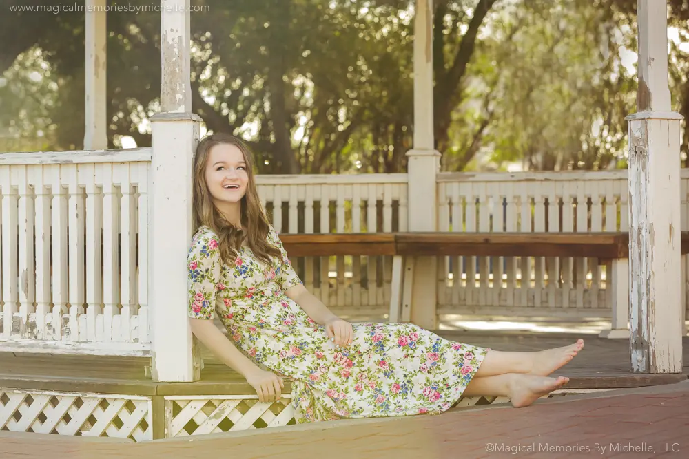 Senior girl lounging barefoot in a floral dress at a white gazebo surrounded by trees in Downtown Mesa.