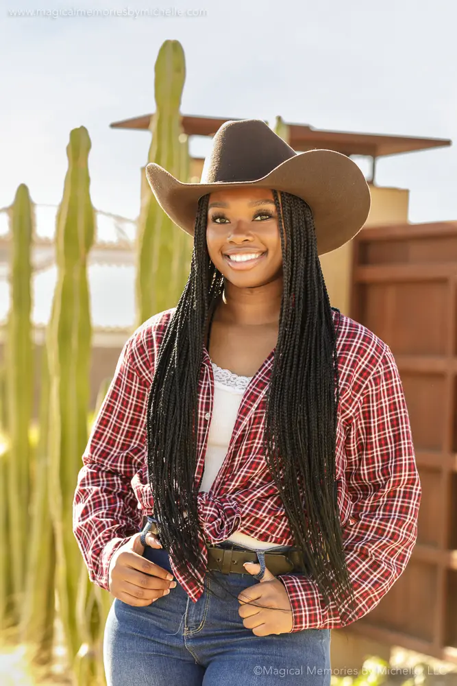 Black high school senior girl wearing a cowboy hat and red plaid shirt smiles in front of a cactus backdrop during a rustic Western-themed senior portrait session in Arizona.