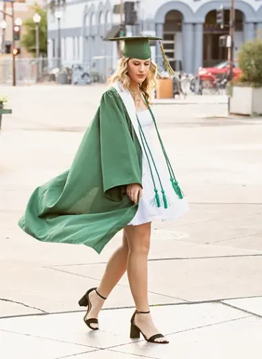 Senior girl in a green cap and gown walking confidently through Downtown Mesa during her senior portrait session.